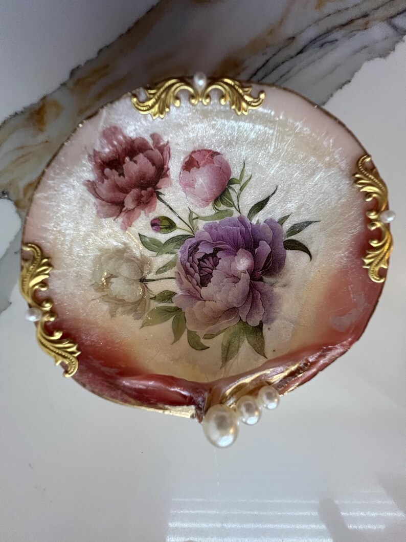 May include: A decorative, shell-shaped dish with a floral design. The dish features a painted arrangement of purple, pink, and white peonies with green leaves. Gold filigree accents the edges, and small pearls are placed around the rim.
