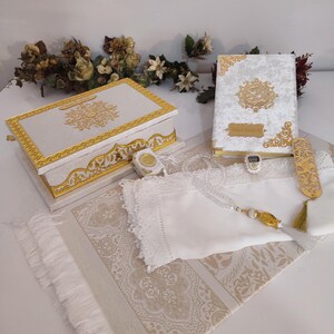May include: A sophisticated set including a white and gold box, a book with gold detailing, a prayer mat, and a white cloth. The box and book feature intricate gold designs. The prayer mat has a beige and white pattern.