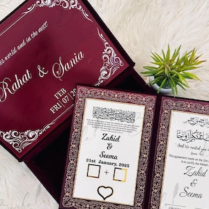 Puede incluir: Caja de invitación de boda de terciopelo burdeos con detalles dorados. La caja abierta revela dos páginas de invitación con marcos dorados y texto. El texto incluye nombres, una fecha y un símbolo de corazón. La tapa de la caja tiene los nombres "Rahat & Sania".