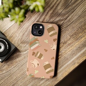 May include: A phone case with a repeating pattern of coffee cups, coffee beans, and stacks of books on a peach background. The case is on a wooden surface with a camera and greenery in the background.