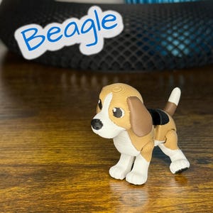 May include: A small, detailed figurine of a beagle dog, featuring tan, white, and black markings. The dog has large, expressive eyes and a white tail tip. The word "Beagle" is written in blue script above the dog.
