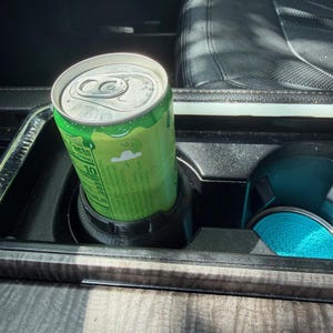 May include: A lime green aluminum beverage can with a silver pull-tab lid sits inside a black plastic cup holder insert. The can features white text and a small cloud graphic, while a bright turquoise circular coaster rests in the adjacent holder.