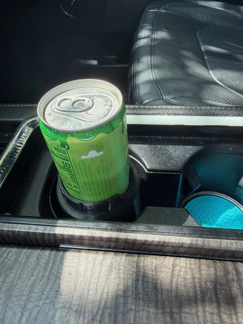 May include: A lime green aluminum beverage can with a silver pull-tab lid sits inside a black plastic cup holder insert. The can features white text and a small cloud graphic, while a bright turquoise circular coaster rests in the adjacent holder.