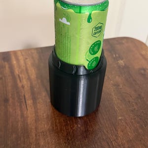 May include: A black cylindrical beverage can cooler holding a green aluminum can. The can features a silver pull tab, a lime graphic, and text including 200mg CAFFEINE, GLUTEN-FREE, and VEGAN. The cooler has a smooth, matte finish and a scalloped top edge.