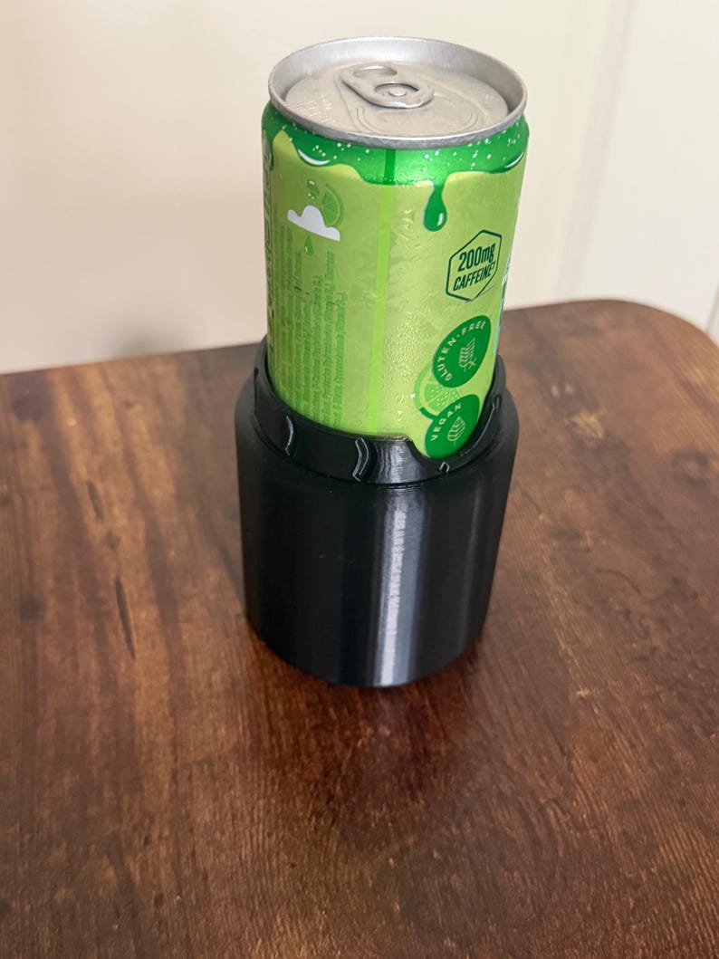 May include: A black cylindrical beverage can cooler holding a green aluminum can. The can features a silver pull tab, a lime graphic, and text including 200mg CAFFEINE, GLUTEN-FREE, and VEGAN. The cooler has a smooth, matte finish and a scalloped top edge.