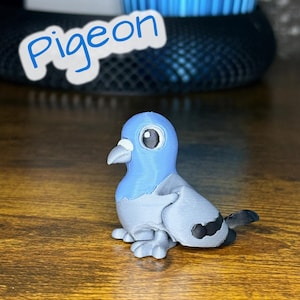 Puede incluir: Una pequeña figura de paloma impresa en tres dimensiones con cabeza azul claro, cuerpo gris y marcas negras en las alas. El ave tiene ojos grandes y redondos y una mancha blanca sobre el pico. La palabra Pigeon está impresa en azul.
