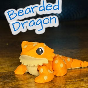 May include: An orange and cream-colored bearded dragon figurine with white spots. The figurine has large black eyes and a frilled neck. The text "Bearded Dragon" is in blue, outlined in white, above the figurine.
