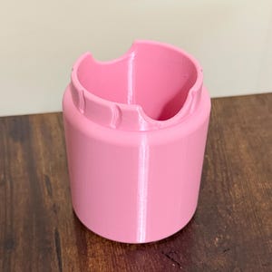 May include: A cylindrical pink plastic holder with a smooth finish. The top edge features a distinct cutout design with two curved indentations, creating a scalloped appearance around the rim of the container. The object has a solid base.