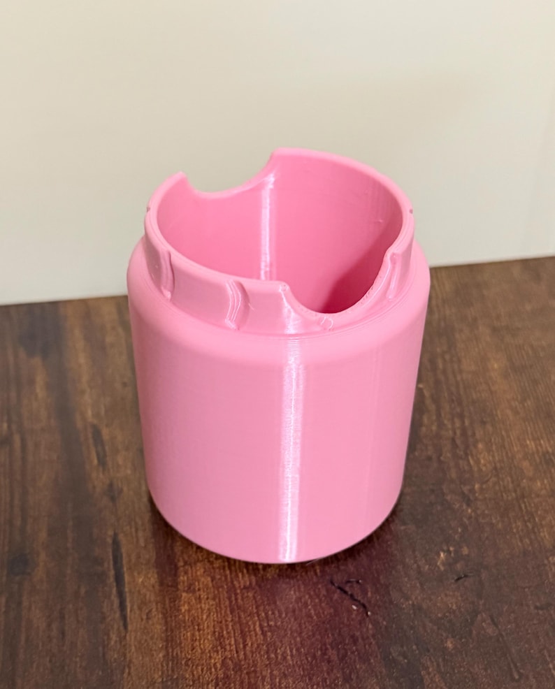 May include: A cylindrical pink plastic holder with a smooth finish. The top edge features a distinct cutout design with two curved indentations, creating a scalloped appearance around the rim of the container. The object has a solid base.