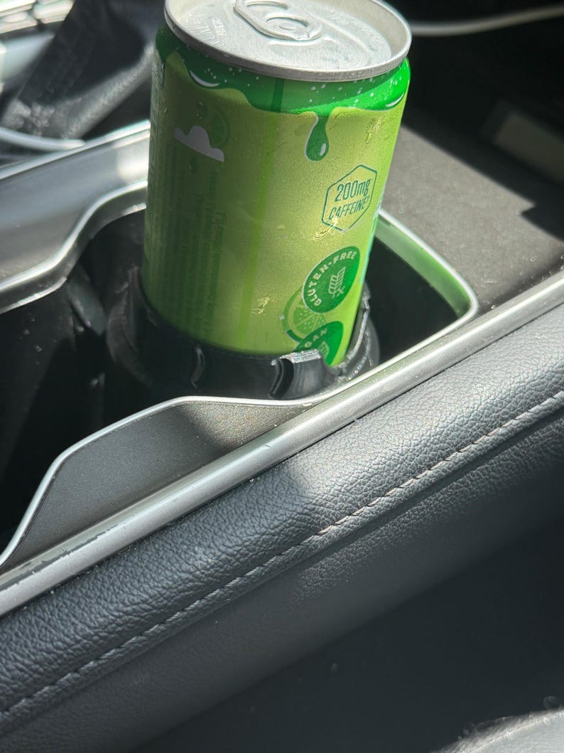 May include: A bright green aluminum beverage can with a silver top sits inside a black plastic cup holder. The can features text that reads 200mg CAFFEINE and GLUTEN-FREE, along with a graphic of a lime slice on the lower portion of the can.