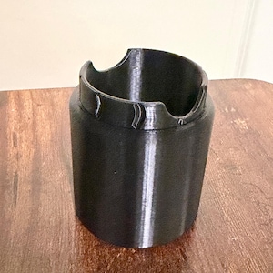May include: A black cylindrical plastic holder with a smooth finish. The top edge features a distinct design with two opposing semi-circular cutouts and raised, curved ridges positioned between the notches, creating a functional rim structure.