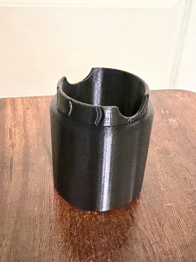 May include: A black cylindrical plastic holder with a smooth finish. The top edge features a distinct design with two opposing semi-circular cutouts and raised, curved ridges positioned between the notches, creating a functional rim structure.