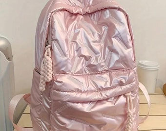 May include: A light pink backpack with a quilted design. The backpack has a front pocket, a top handle, and adjustable shoulder straps. A decorative tassel hangs from the zipper. The bag is made of a shiny, reflective material.