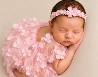 May include: A light pink newborn outfit featuring a sheer tulle dress adorned with numerous small fabric flowers and a matching headband with similar floral embellishments. The garment is designed with a soft, layered texture and delicate petals.