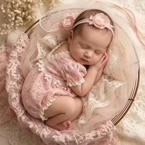 May include: A sleeping infant wearing a pale pink lace romper with short puff sleeves and white floral trim, paired with a matching headband featuring a soft pink fabric flower. The baby is nestled within a round, cream-colored lace-lined basket.