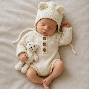 May include: A cream-colored knitted long-sleeve romper featuring three brown wooden buttons on the front placket, paired with a matching bear-ear bonnet. A small, cream-colored crocheted teddy bear is held in one hand, completing the set.