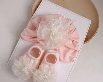 May include: A baby's pink turban and socks set. The turban features a large white flower with lace detailing. The socks have a matching white flower accent. The set is displayed on a white card with the brand name "Chibaby".