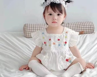 Puede incluir: Un body blanco para bebé con bordados florales coloridos, con mangas con volantes y un escote cuadrado. El conjunto se combina con calcetines altos blancos hasta la rodilla y una diadema de encaje. El body está hecho de una tela suave y texturizada.