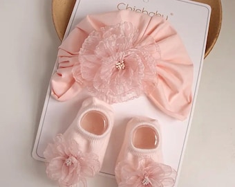 May include: A baby set featuring a pink turban-style hat with a large, layered flower and a pair of matching socks. The socks and hat have a delicate, feminine design. The text "Chichaby" is visible on the packaging.