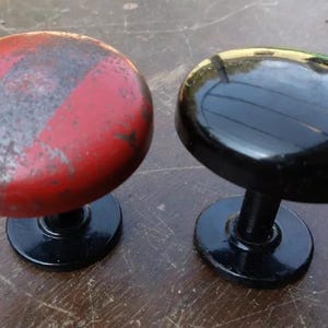 以下が含まれることがあります： 2つのヴィンテージドアノブ。1つは使用感のある赤色、もう1つは黒色で光沢があります。どちらも丸いベースとステムが付いています。ノブはおそらく金属製です。