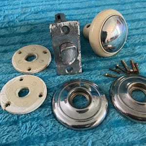 May include: A collection of vintage door hardware, including a chrome doorknob with a cream-colored base, a metal latch, two chrome escutcheons, two white oval plates, and several screws. The hardware is arranged on a blue textured surface.