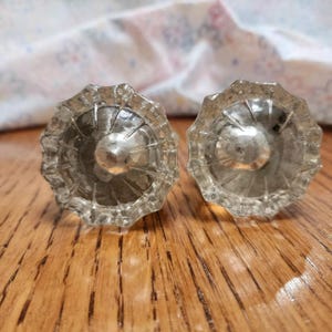 May include: A pair of clear glass cabinet knobs with a faceted design. The knobs feature a round, slightly domed centre and a scalloped edge. They are set against a wooden surface with a floral patterned fabric in the background.