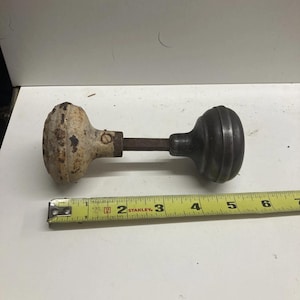 May include: An antique door knob set with a white and black finish. The set includes two knobs connected by a metal rod. A yellow measuring tape is next to the door knob set, measuring approximately 7 inches.