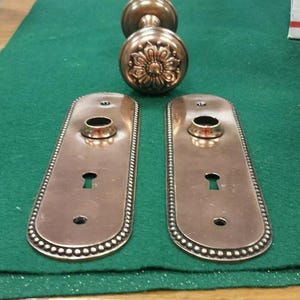 Puede incluir: Juego de herrajes de puerta de bronce antiguo. El juego incluye dos placas traseras rectangulares con cerraduras y un borde decorativo con cuentas, y dos pomos a juego. Un pomo presenta un diseño floral.