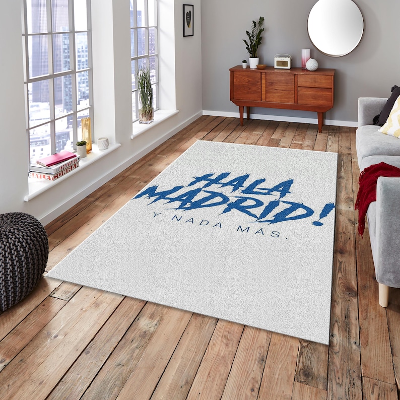 May include: White rectangular area rug with the words "Hala Madrid!" in blue script. The rug is placed on a hardwood floor in a room with a window and a wooden cabinet.