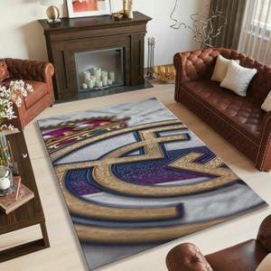 May include: A rectangular area rug featuring a large, centered Real Madrid football club logo. The design displays a gold crown, a blue and gold circular emblem with the letters MFC, and a diagonal purple stripe, all set against a textured gray background.