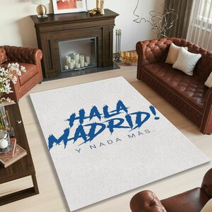 May include: White rectangular rug with the text "HALA MADRID! Y NADA M&Aacute;S" in blue. The rug is placed on a light-colored wooden floor in a living room setting. Brown leather sofas and a fireplace are visible in the background.