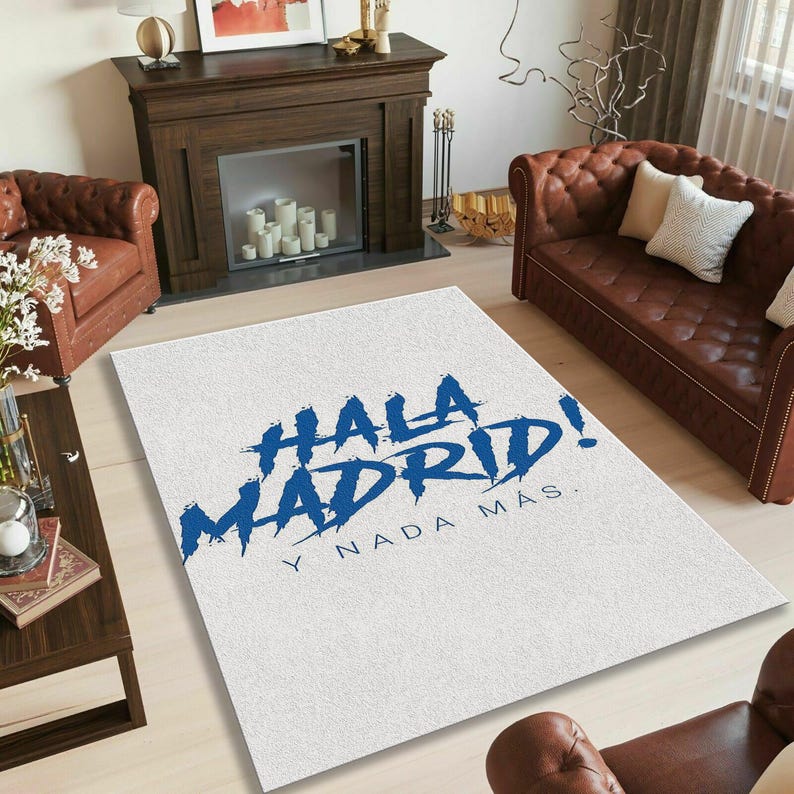 May include: White rectangular rug with the text "HALA MADRID! Y NADA M&Aacute;S" in blue. The rug is placed on a light-colored wooden floor in a living room setting. Brown leather sofas and a fireplace are visible in the background.