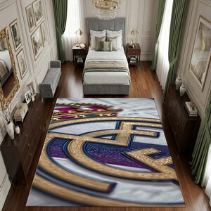 May include: A rectangular area rug featuring a large, centered Real Madrid soccer club logo. The design displays a gold crown, a blue and gold circular emblem, and a white background. The rug is positioned on a wooden floor in a bedroom.