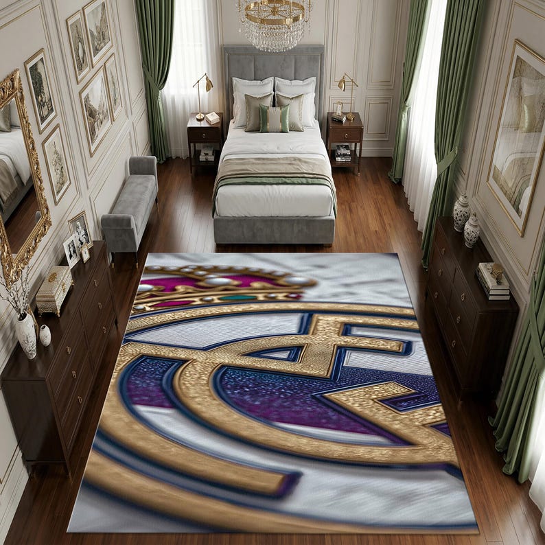 May include: A rectangular area rug featuring a large, centered Real Madrid soccer club logo. The design displays a gold crown, a blue and gold circular emblem, and a white background. The rug is positioned on a wooden floor in a bedroom.