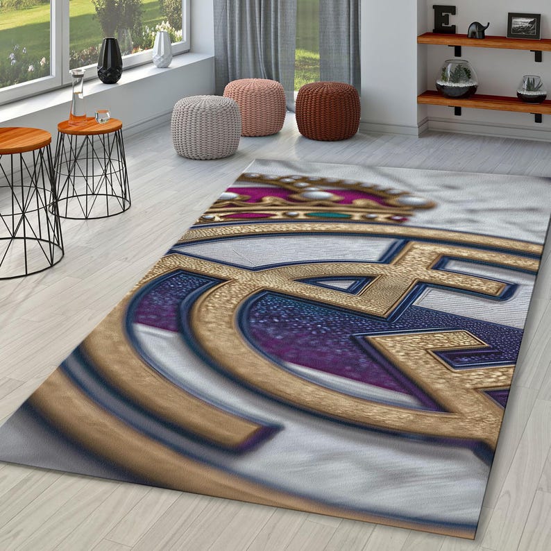 May include: A rectangular area rug featuring a large, centered Real Madrid football club logo. The design includes a gold crown with blue and red accents, set against a white background with a textured, metallic-looking finish on the emblem.
