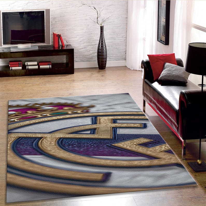 May include: A rectangular area rug featuring a large, detailed graphic of the Real Madrid football club crest. The design displays a gold crown, a blue diagonal stripe, and the intertwined letters MCF in gold, set against a white background.