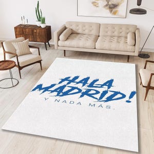 May include: A white rectangular area rug with the words "HALA MADRID!" in blue, along with "Y NADA M&Aacute;S." The rug is in a living room setting with a beige sofa, wooden furniture, and a wooden floor.