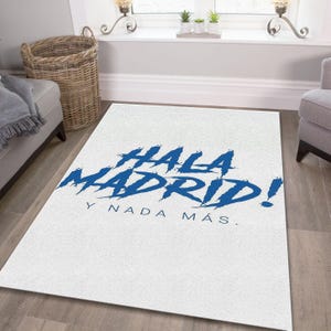 May include: White rectangular rug with the words "HALA MADRID!" in blue brush lettering. Below the text is "Y NADA M&Aacute;S." The rug is on a hardwood floor, with a wicker basket and a gray sofa in the background.