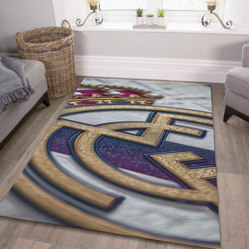 May include: A rectangular area rug featuring a large, stylized gold and blue emblem with a crown at the top. The design includes textured gold lines, deep blue accents, and a white background, creating a bold graphic pattern across the floor covering.