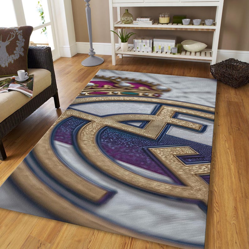 May include: A rectangular area rug featuring a large, stylized gold and blue emblem with a crown at the top. The design includes a textured gold metallic-look border, a deep blue central section, and a white background with a subtle brushstroke texture.