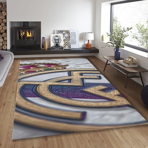 May include: A rectangular area rug featuring a large, centered Real Madrid soccer club logo. The design displays a gold crown at the top, with gold, blue, and white circular elements forming the emblem against a textured, light-colored background.