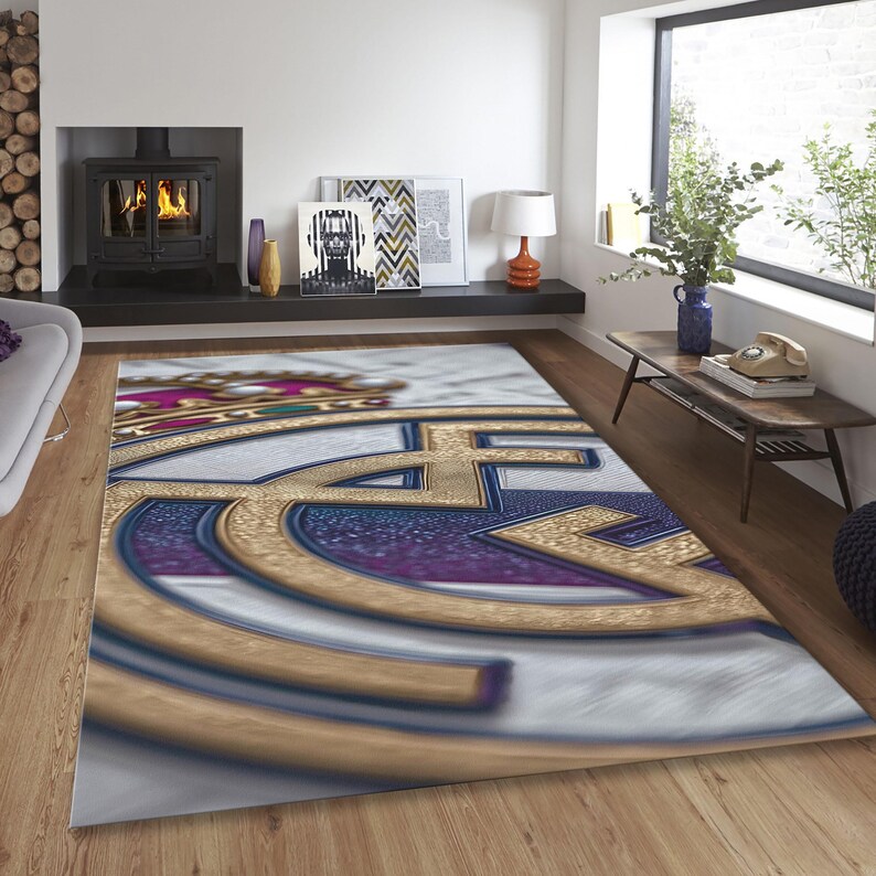 May include: A rectangular area rug featuring a large, centered Real Madrid soccer club logo. The design displays a gold crown at the top, with gold, blue, and white circular elements forming the emblem against a textured, light-colored background.