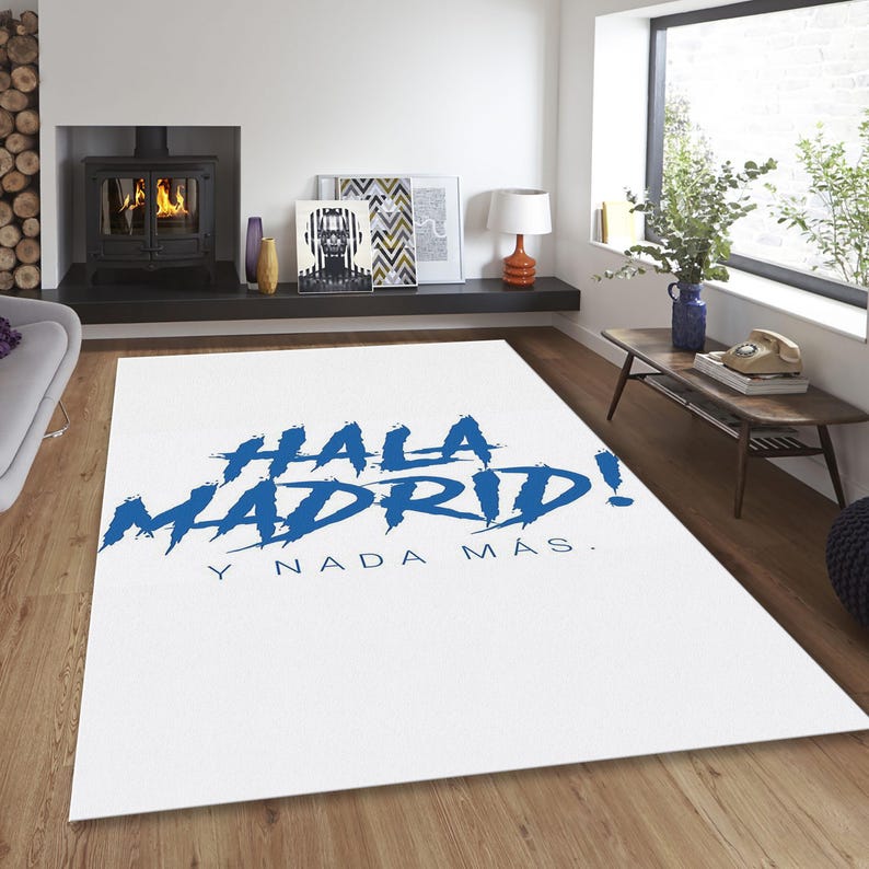 May include: White rectangular rug with the words "HALA MADRID!" in blue, and "Y NADA M&Aacute;S." below. The rug is in a room with a fireplace, artwork, and a wooden table.