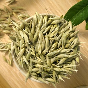 May include: A white ceramic bowl filled with dried oat grains, set on a light wooden tray. The grains are a pale green color, with long, pointed shapes. Some grains are scattered around the bowl.