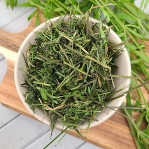 May include: A white ceramic bowl filled with dried green herbs, possibly for culinary use. The bowl sits on a wooden cutting board, with fresh green sprigs of the same herb scattered around. The herbs have a mix of green and brown hues.