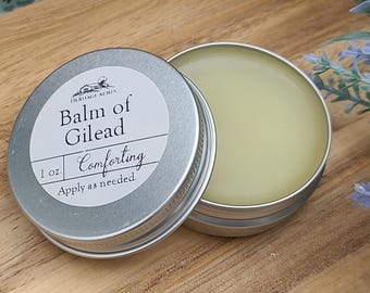 May include: A round silver metal tin containing a pale yellow balm, with a white label on the lid reading Heritage Acres Balm of Gilead Comforting 1 oz. Apply as needed. The lid is placed beside the open tin, revealing the smooth balm inside.
