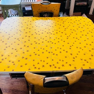 May include: A retro yellow dining table with a starburst design, accompanied by matching chairs. The table features a chrome trim and sits on a dark wooden floor. The chairs have yellow upholstery and decorative backs.