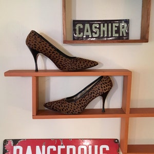 Op de afbeelding: Twee pumps met luipaardprint met zwarte hakken en afwerking worden tentoongesteld op een oranje plank. Boven de schoenen staat een vintage metalen bord met de tekst "CASHIER". Onder de schoenen staat een rood en wit bord met de tekst "DANGEROUS".