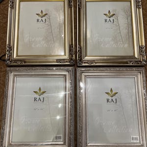 May include: Four silver picture frames with ornate detailing. Each frame features a white mat and a clear glass front. The frames are all the same size and have the text "RAJ Frame Collection" printed on the mat. The frames are 11 inches by 14 inches.