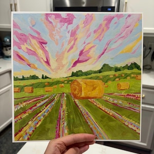 Puede incluir: Una impresi&oacute;n art&iacute;stica cuadrada que muestra un paisaje con un campo verde, varias pacas de heno doradas y un cielo vibrante lleno de nubes con tonos rosas, amarillos y azul claro. El primer plano muestra filas de flores coloridas.